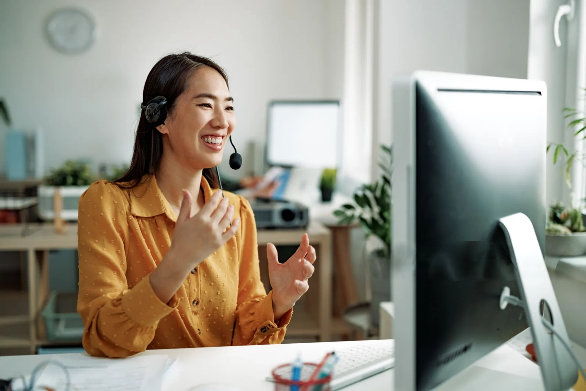 Smiling Asian customer service representative in a yellow blouse wearing a headset, gesturing during a video call in a bright, modern office space.