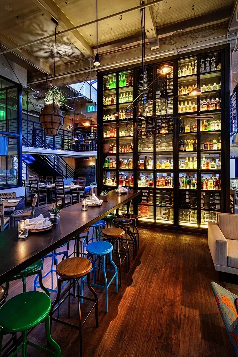 Modern industrial bar with warm ambient lighting, colorful stools, and a towering, backlit grid display of spirits, featuring an "EXIT" sign above.