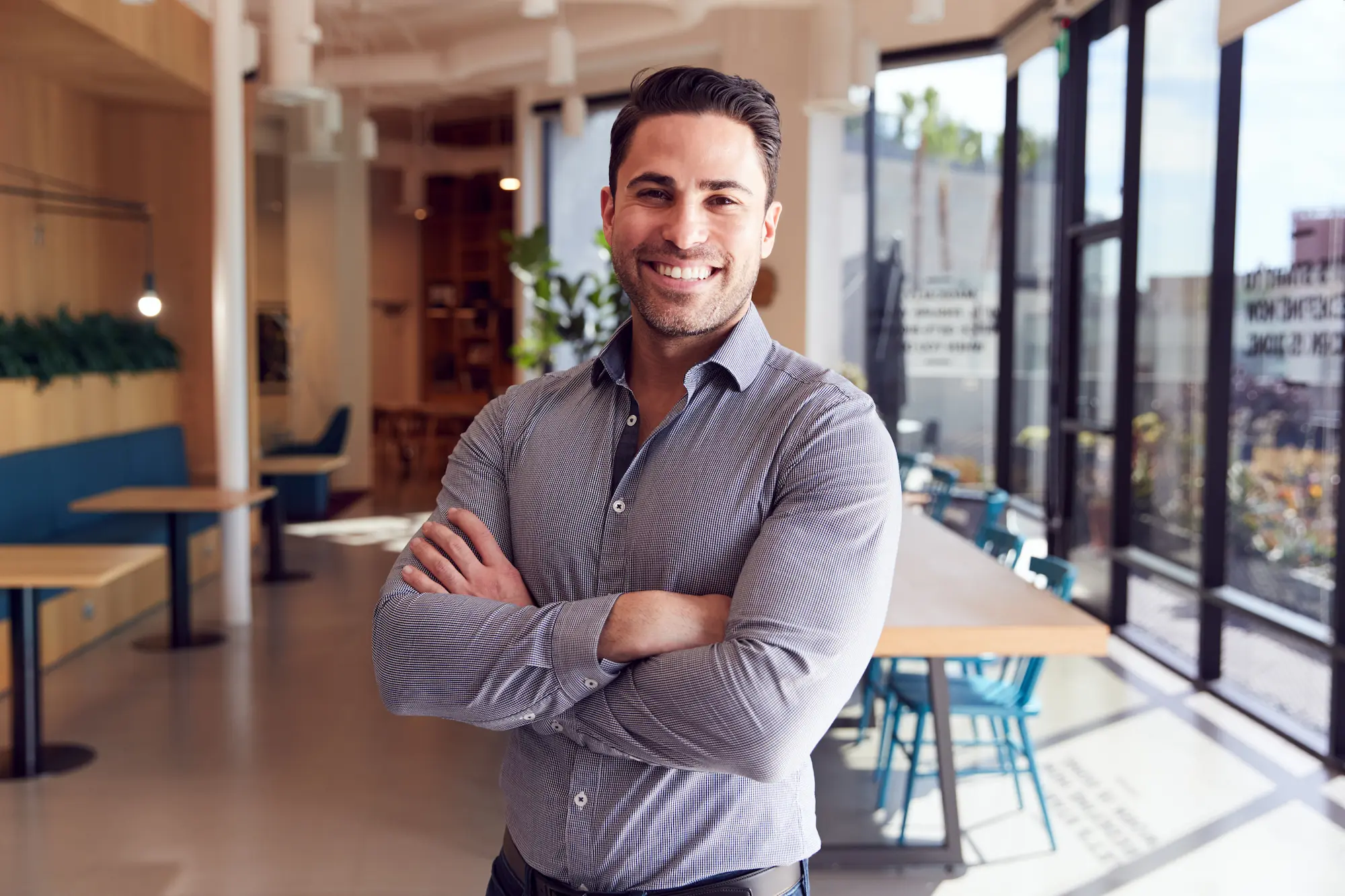 Smiling man in dark blue shirt, arms crossed, standing in a brightly lit modern office. Background shows blurred coworkers.