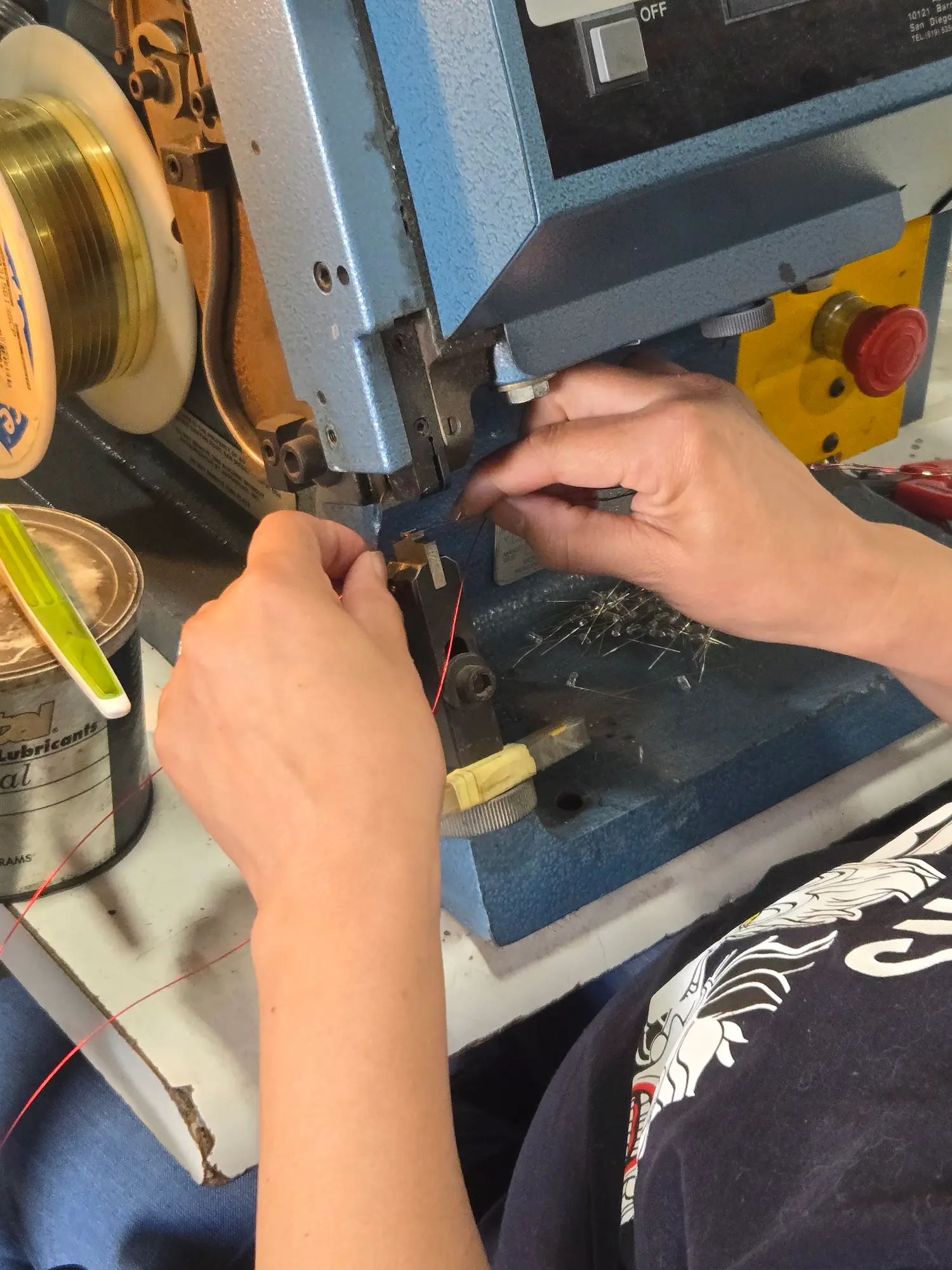 About-Facility Person operates a blue industrial machine, feeding a wire into it. Spool of wire, lubricant can, and yellow emergency stop button are visible nearby.