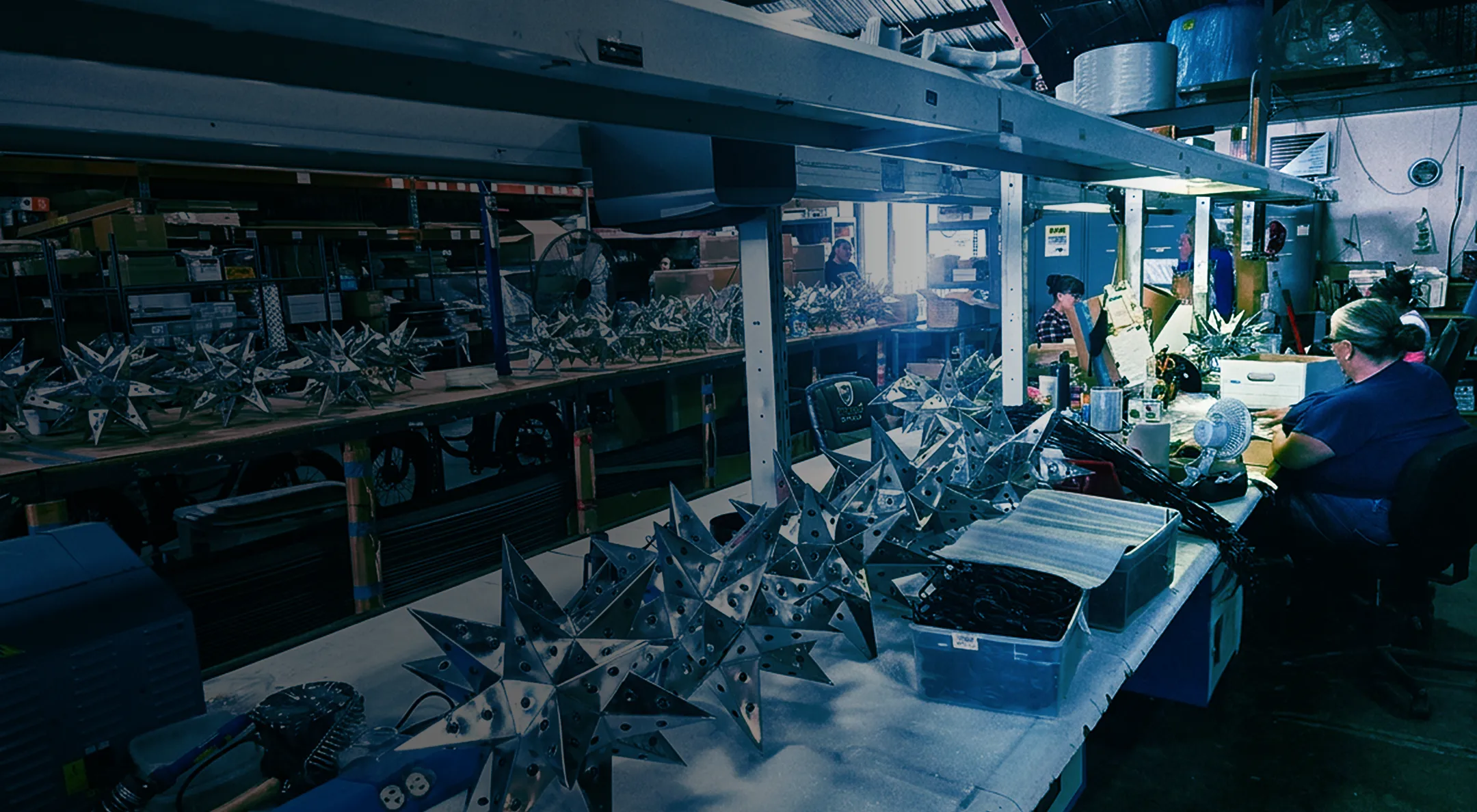 About-Hero-Gradient Busy metalworking workshop with rows of shiny star-shaped sculptures on tables and shelves, while workers assemble pieces in the background.
